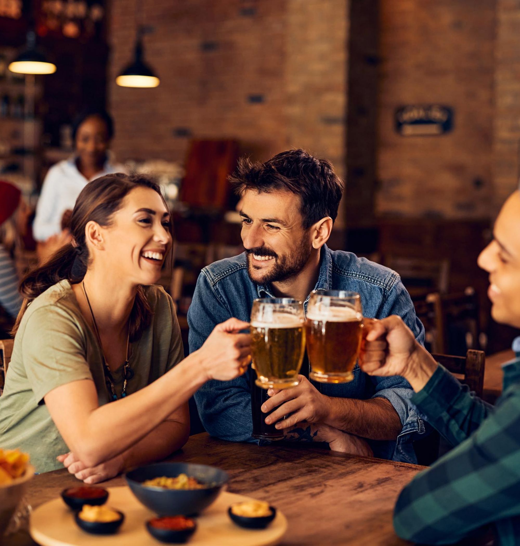 Three friends toast with beer mugs at a cozy pub, smiling and enjoying appetizers at a wooden table near LOCAL Boise apartments.