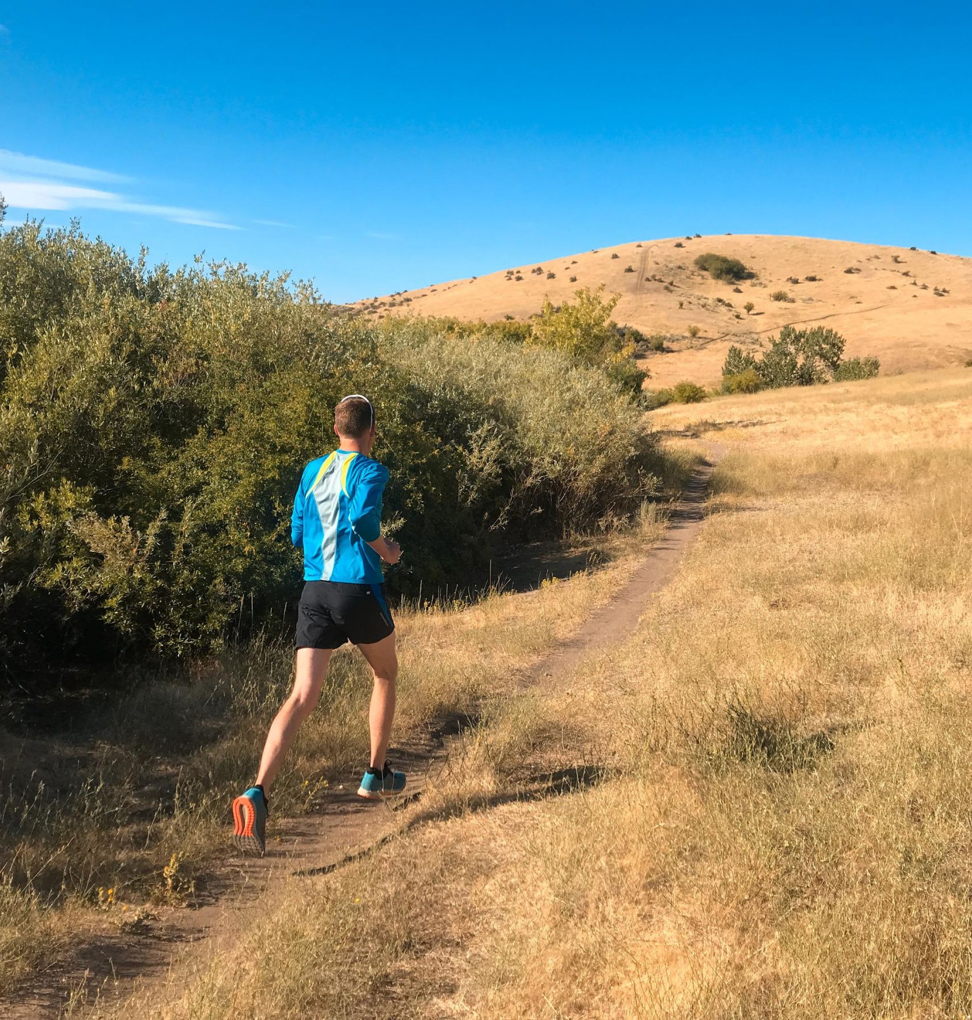 Person running on a trail through a dry, grassy landscape with a hill under a clear blue sky near LOCAL Boise apartments.