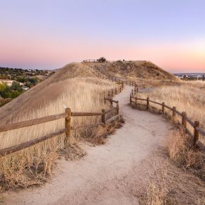 Dirt path lined with wooden fences winding through dry grassy hills at sunset, with a city in the distance near LOCAL Boise apartments.