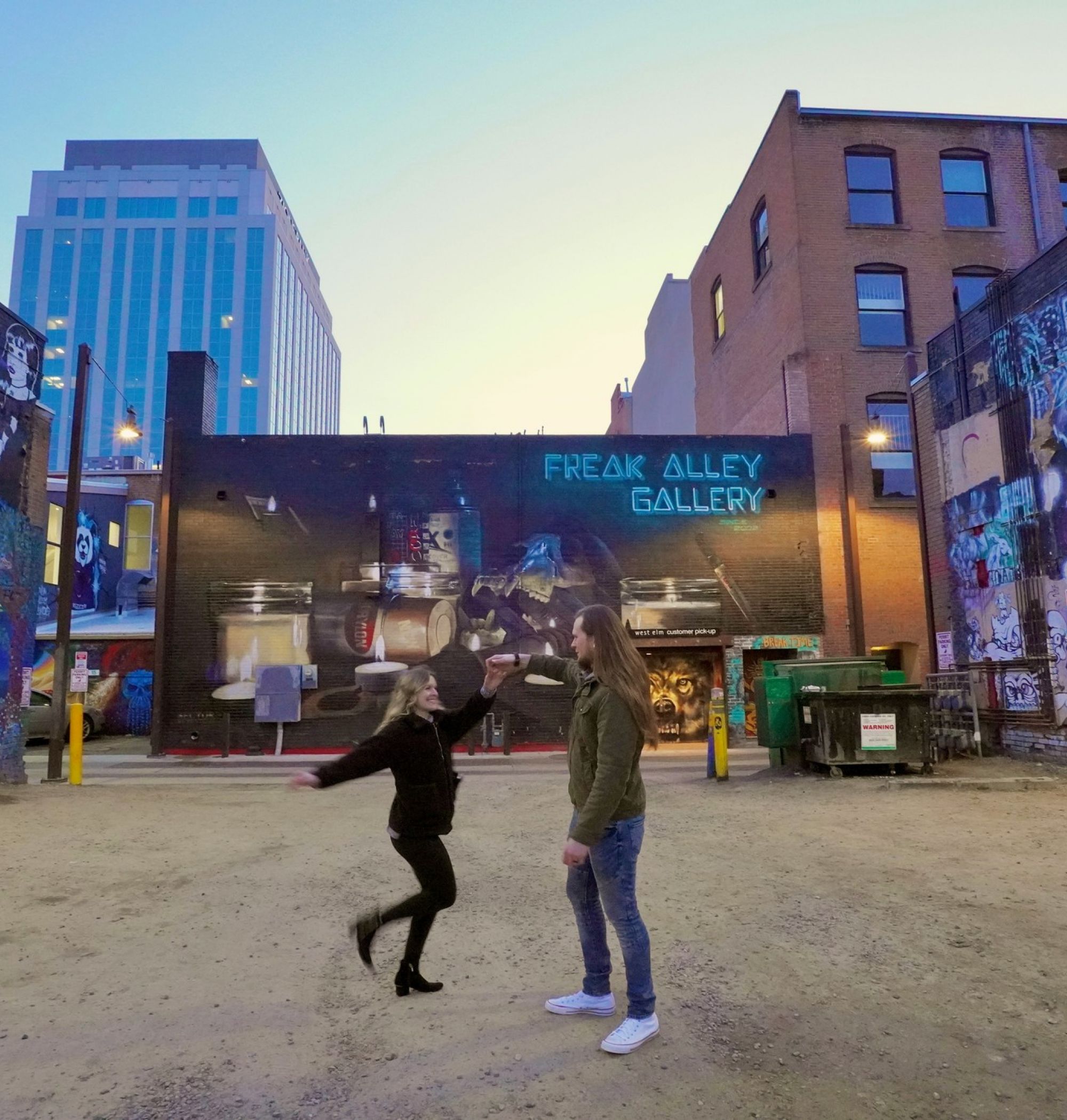 Two people stand in a graffiti-filled alley with "Freak Alley Gallery" written on the wall at dusk near LOCAL Boise apartments.