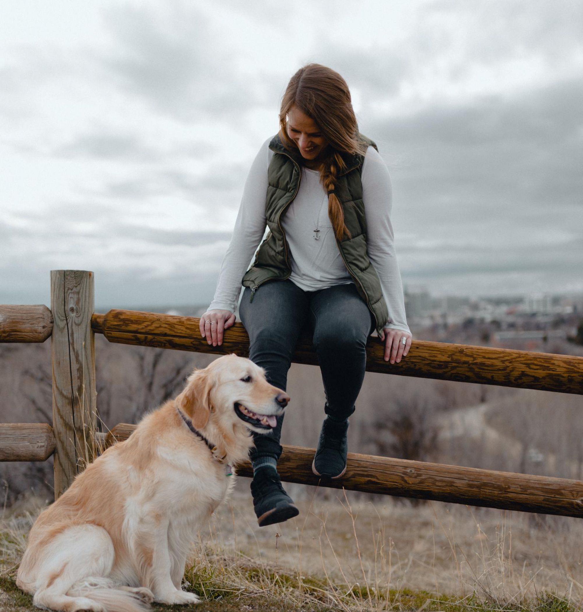 A person sitting on a wooden fence with a golden retriever nearby on a grassy hill, under a cloudy sky near LOCAL Boise apartments.